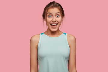 Fototapeta premium Joyful woman with bugged eyes, has toothy smile, dressed in casual vest, isolated over pink background, enjoys free time, hears something unexpected and pleasant. Reaction and people concept
