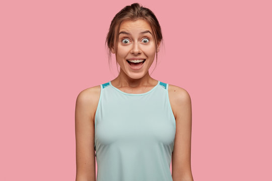 Joyful woman with bugged eyes, has toothy smile, dressed in casual vest, isolated over pink background, enjoys free time, hears something unexpected and pleasant. Reaction and people concept