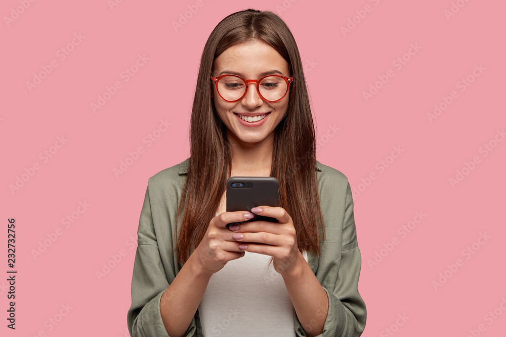 Wall mural satisfied lovely woman holds modern cell phone, texts with friend online in chat, dressed in stylish