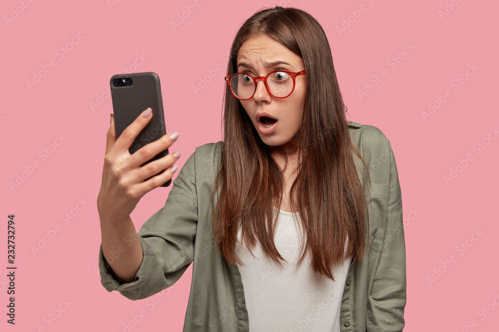Wall mural shocked woman sees embarrasing picture in screen of smart phone, stares with amazement at camera, we - Wall murals