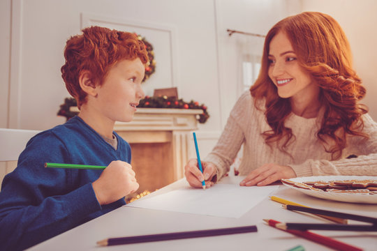 Quality time together. Pleasant red-haired preteen boy and his young mother sitting at the table and drawing a gingerbread man together while smiling at each other