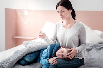 Temperature raising up. Portrait of young worried mother sitting on the bed while looking at thermometer and keeping her daughter on her knees