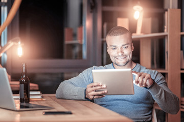 Deep at night. Attractive international programmer expressing positivity and looking at tablet while typing message