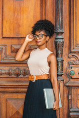 Young African American College Student with afro hairstyle, eye glasses, wearing sleeveless light color top, black skirt, belt, holding laptop computer, standing by vintage office door in New York..
