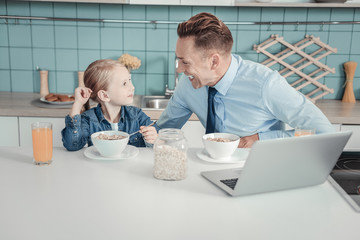 Best father. Lovely attentive occupied father sitting by the table in the kitchen having breakfast and smiling to his daughter.