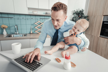Little helper. Pleasant busy concentrated man standing in the kitchen hugging a nursling who sitting on the table and using the laptop.
