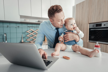 Im with you. Pleasant lovely attentive father spending time in the kitchen looking at baby and using the laptop.