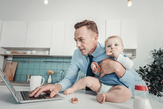 Family business. Occupied caring handsome man standing in the kitchen near the table hugging a baby and working with the laptop.