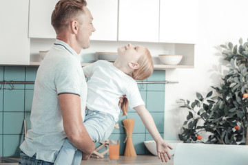 Play with me. Happy attentive pleasant father standing in the bright room holding daughter and...