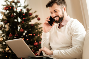 Distance communication. Delighted positive handsome man smiling and making a call while looking at the laptop screen