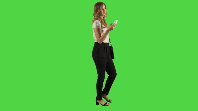 A young businesswoman walking and texting gladly while looking into the camera in a full body shot over a green screen.