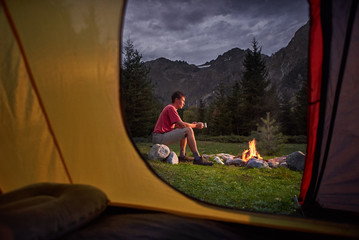 View from inside tourist tent at sunset. Alone tourist, man hiker sitting at bonfire