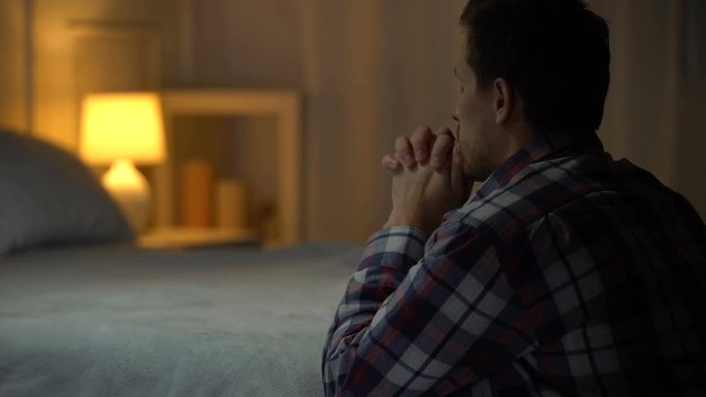Religious young man praying in evening near bed, belief in God, Christianity