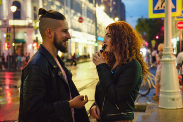 Couple in love on date. Boyfriend talking to girlfriend. She smokes an electronic cigarette. They walk streets of night city. Wet asphalt after rain.