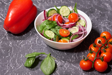 Vegetable salad in a white plate on a gray background, next to the Bulgarian pepper, tomatoes, spinach. Selective focus