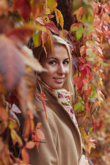 Authentic vertical close up of young Caucasian adorable blond woman with deep blue eyes, mysteriously looking at camera, hiding in huge wall from autumn leaves, posing on colorful natural background.