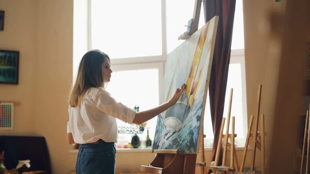 Busy young woman is casual clothing is painting with oil paints standing near easel and holding brush and palette. Hobby, art and creative people concept.