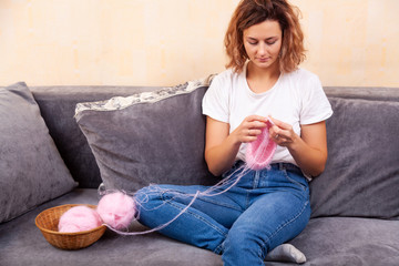 Fototapeta premium Close up of a young married woman dressed in trendy d jeans and t-shirt knitting a pink hat with knitting needles in home. Freelance creative working and living concept