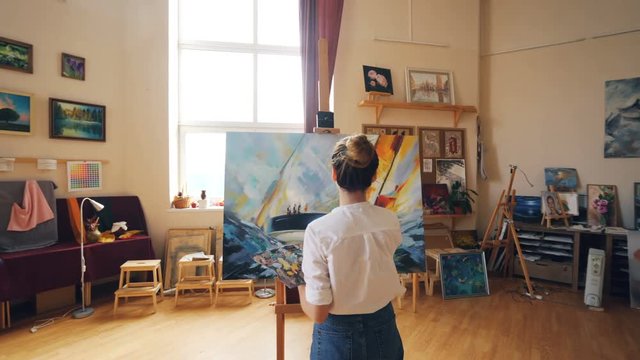 Pretty blond girl artist is painting marine landscape working in light workshop alone using brush and palette. Tools and artworks on shelves are visible.