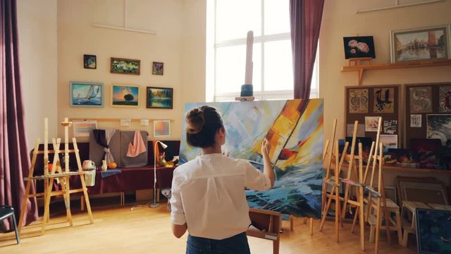 Pan shot of young woman focused on painting seascape on canvas in nice light studio standing near easel alone and working. Fine arts and people concept.