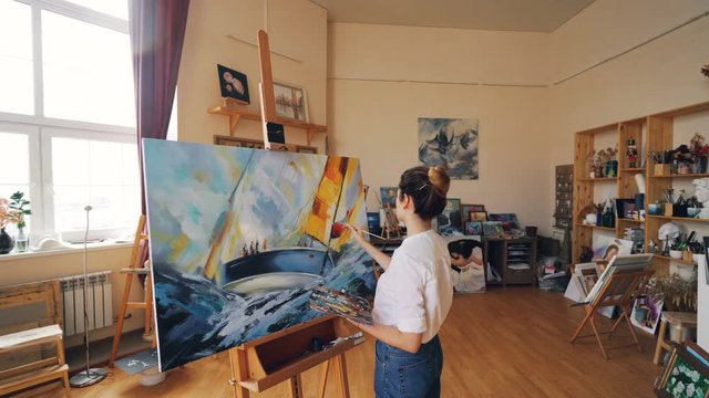 Pan shot of female painter depicting marine landscape on canvas using oil paints, brush and palette working in workshop alone. Visual arts and youth concept.