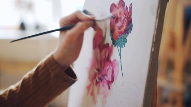 Close-up shot of female hand holding paintbrush and painting bright flowers on canvas in light studio. Pictorial art, artistic work and people concept.