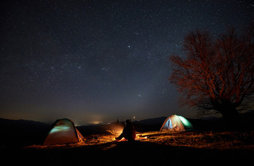 Night camping in mountains. Silhouette of male tourist having a rest between two illuminated tents, enjoying campfire, beautiful dark blue starry sky on background. Tourism, outdoor activity concept