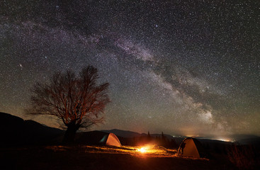Amazing night camping site view. Bright campfire burning between two tourist tents under incredible beautiful starry sky and Milky way. Big tree and distant mountain range on background
