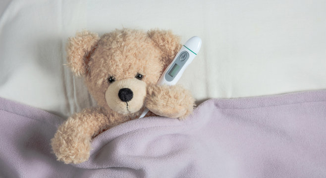 Cold, flu or allergy. Cute teddy in bed, covered with a warm blanket, holding a tissue