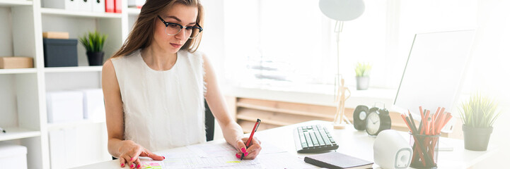 Beautiful young girl is sitting at the table in the office, holding a pen in her hand and filling...