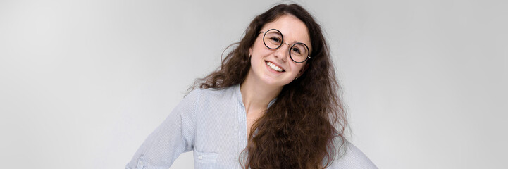 Obraz premium Portrait of a happy young girl in glasses. Beautiful young girl on a gray background. A young girl in a light blouse and dark trousers.