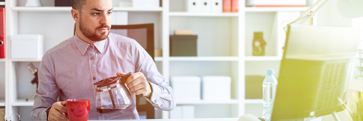A young man in the office sits at a table, looks at the monitor and pours coffee into a cup.