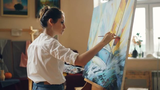 Professional artist young woman is painting seascape with acrylic paints finishing marine landscape ship and sea waves working alone in cozy studio. People and work concept.