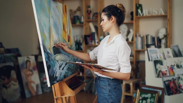 Cheerful young woman artist is painting beautiful picture marine landscape using oil paints then looking at masterpiece and smiling enjoying her work.
