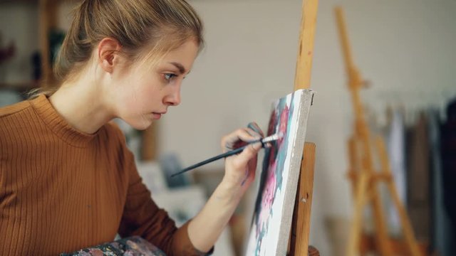 Diligent art student pretty girl is painting beautiful picture flowers working in class standing near easel. Person is concentrated on creating artwork.