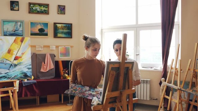 Female art teacher is working with good-looking young girl student painting picture giving advice standing in front of easel with paintbrush and palette.