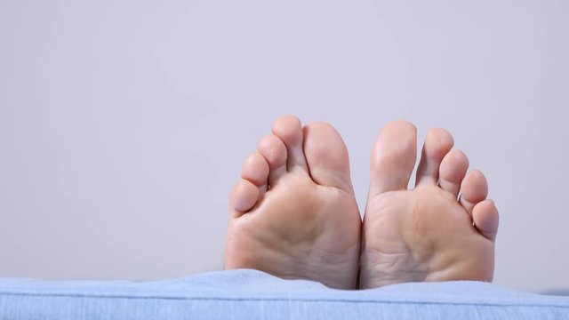 Female resting on sofa showing bare foot. Human feet moving on bed in slow motion, white grey wall background copy space text area