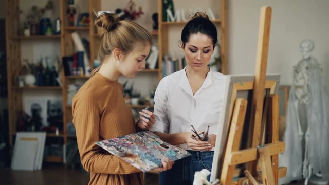 Friendly art teacher good-looking woman in casual clothing is teaching female student talking then giving her brush, girl is smiling and mixing paints on palette.