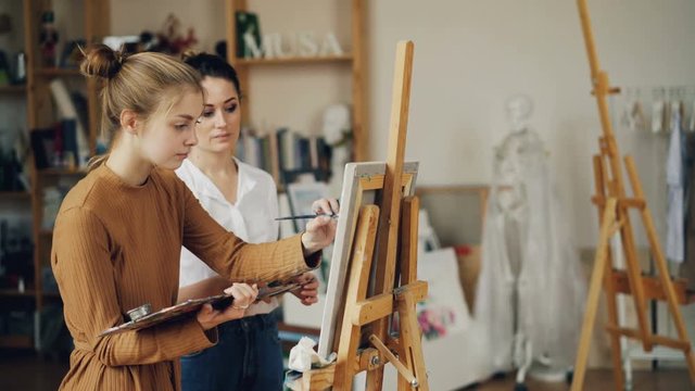 Cute teenage girl is learning painting from experienced teacher in art school in workroom with artworks and wooden easels. Artist is teaching talking and pointing at picture.