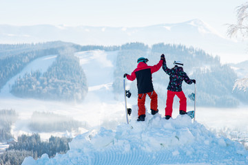 Shot of a couple high fiving each other posing on top of a snowy mountain observing stunning winter...