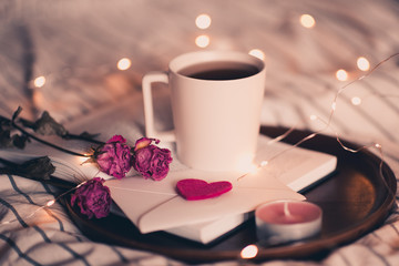 Cup of tea staying on open book with flowers and envelope in bed over lights at background. Valentines day.
