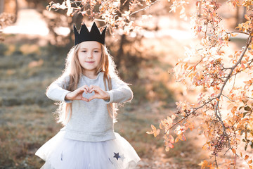 Smiling kid girl 4-5 year old making heart with hands wearing handmade crown outdoors. Looking at...