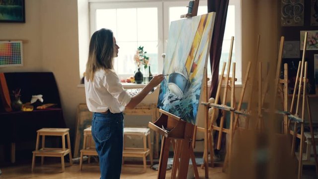 Slender blond girl is painting picture with oil paints holding brush and depicting seascape working indoors in workroom. Artistry and creative people concept.
