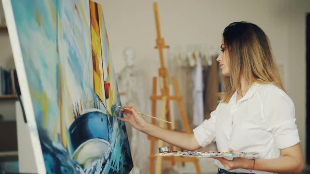 Blond girl painter is working in studio depicting sea landscape and boat on canvas using brush and tempera paints looking at picture and enjoying her occupation.