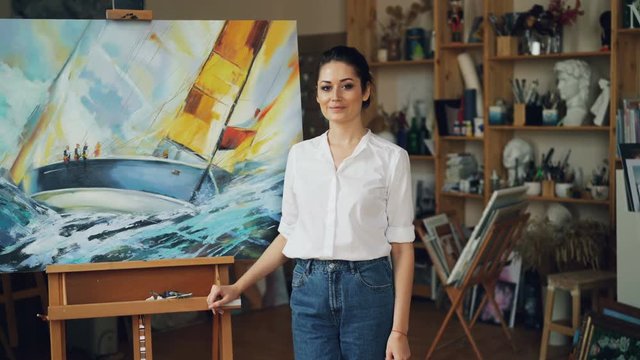 Portrait of pretty young lady painter looking at camera and smiling standing near her beautiful picture on easel in modern studio enjoying her work.