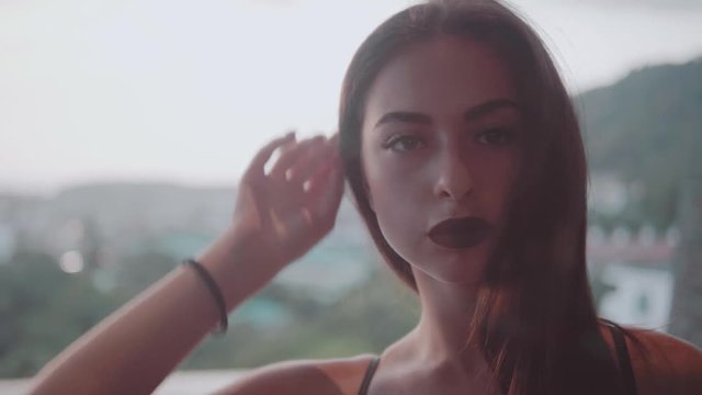 Portrait of sexy brunette woman in black bra posing outdoor over cloudy sky background. View from inside of apartment through the terrace window - video in slow motion
