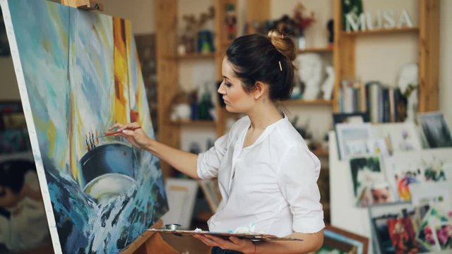 Attractive woman painter is painting ship and sea on picture creating beautiful seascape using acrylic paints working in traditional technique in studio.