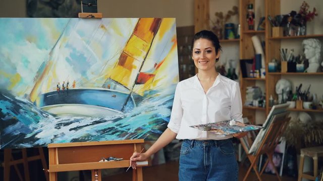 Portrait of attractive young woman painter looking at camera and smiling standing near her beautiful picture in modern studio holding palette and brush.