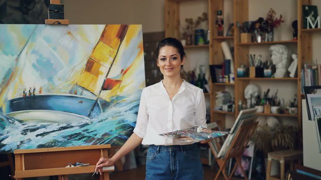 Portrait of confident girl painter holding palette and paintbrush standing near seascape looking at camera and smiling. Modern workshop with artworks is in background.