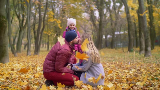 Beautiful parents sitting squatted, kissing tenderly in autumn park while adorable elementary age daughter throwing colorful autumn foliage over them. Happy family relaxing outdoors in fall season.